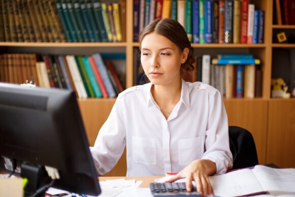 Thoughtful female administrative assistant checking computer reading information report and noting data skilled woman watching online video online thinking over reports writing in notepad